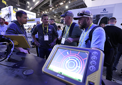 People at CONEXPO-CON/AGG gathered around a booth with electronic displays showing colorful circular graphics, with company logos and banners in the background