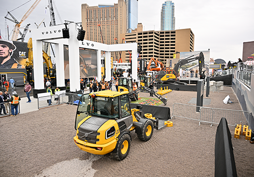 Construction equipment expo with a yellow Volvo wheel loader in front, surrounded by excavators, cranes, and visitors, with banners from Volvo and CAT in the background