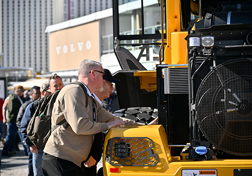 A group of people closely examining VOLVO construction vehicle at CONEXPO-CON/AGG festival lot, with a building labeled 'VOLVO' visible in the background