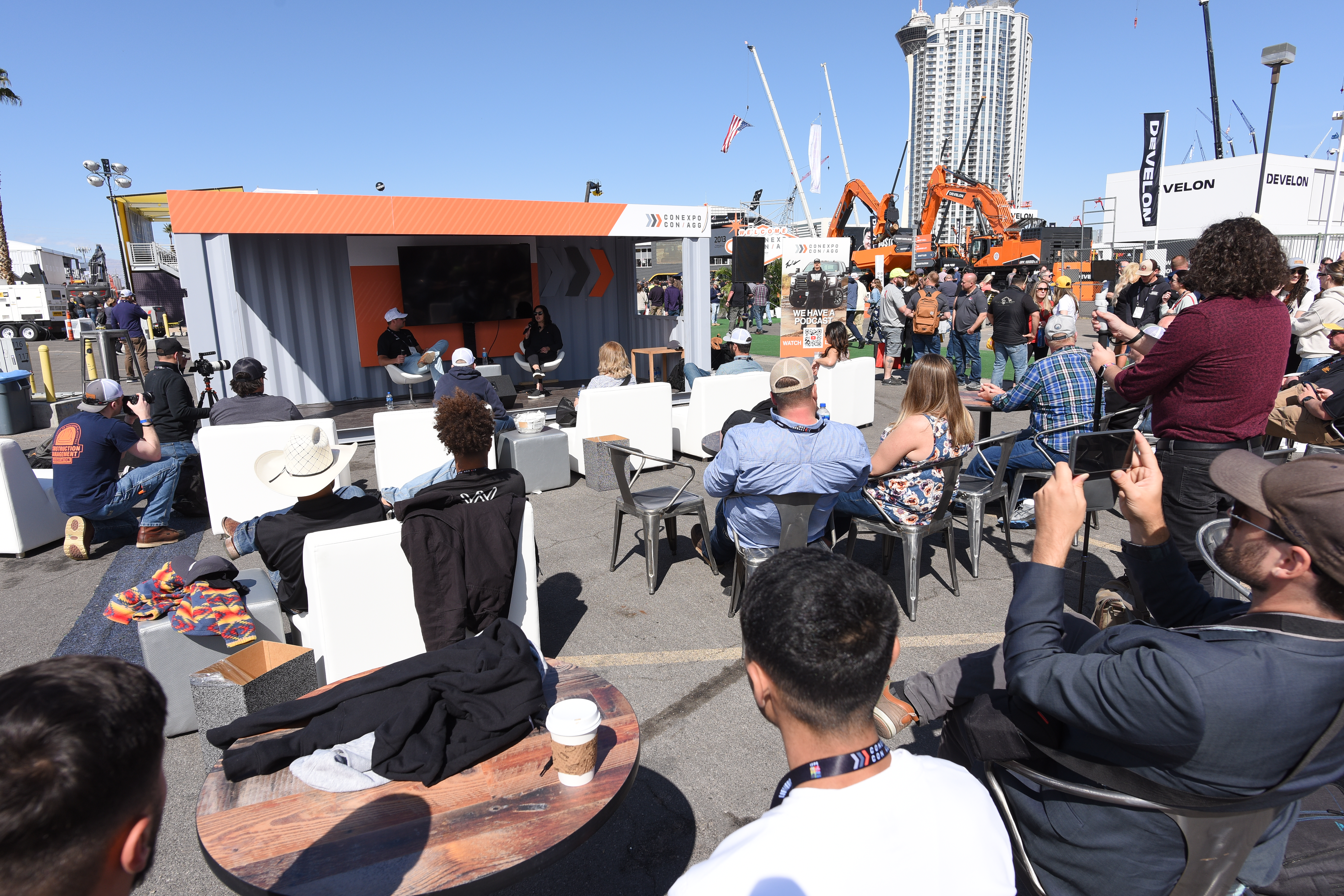Outdoor audience watching a presentation at the Community Zone made from a shipping container, with city buildings and construction equipment in the background