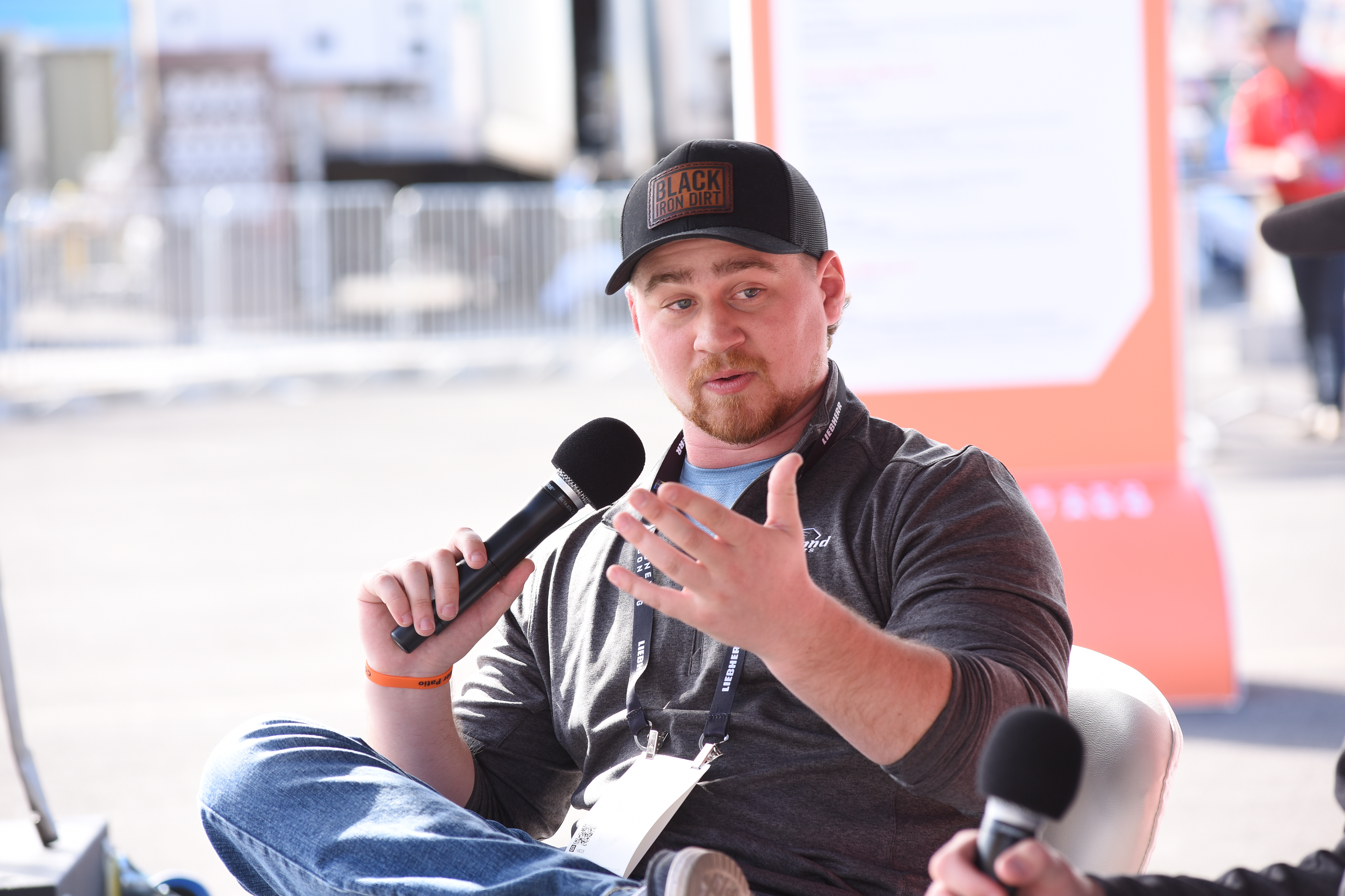 Person wearing a 'BLACK' cap holding a microphone during an interview, with blurred background and another person also holding a mic
