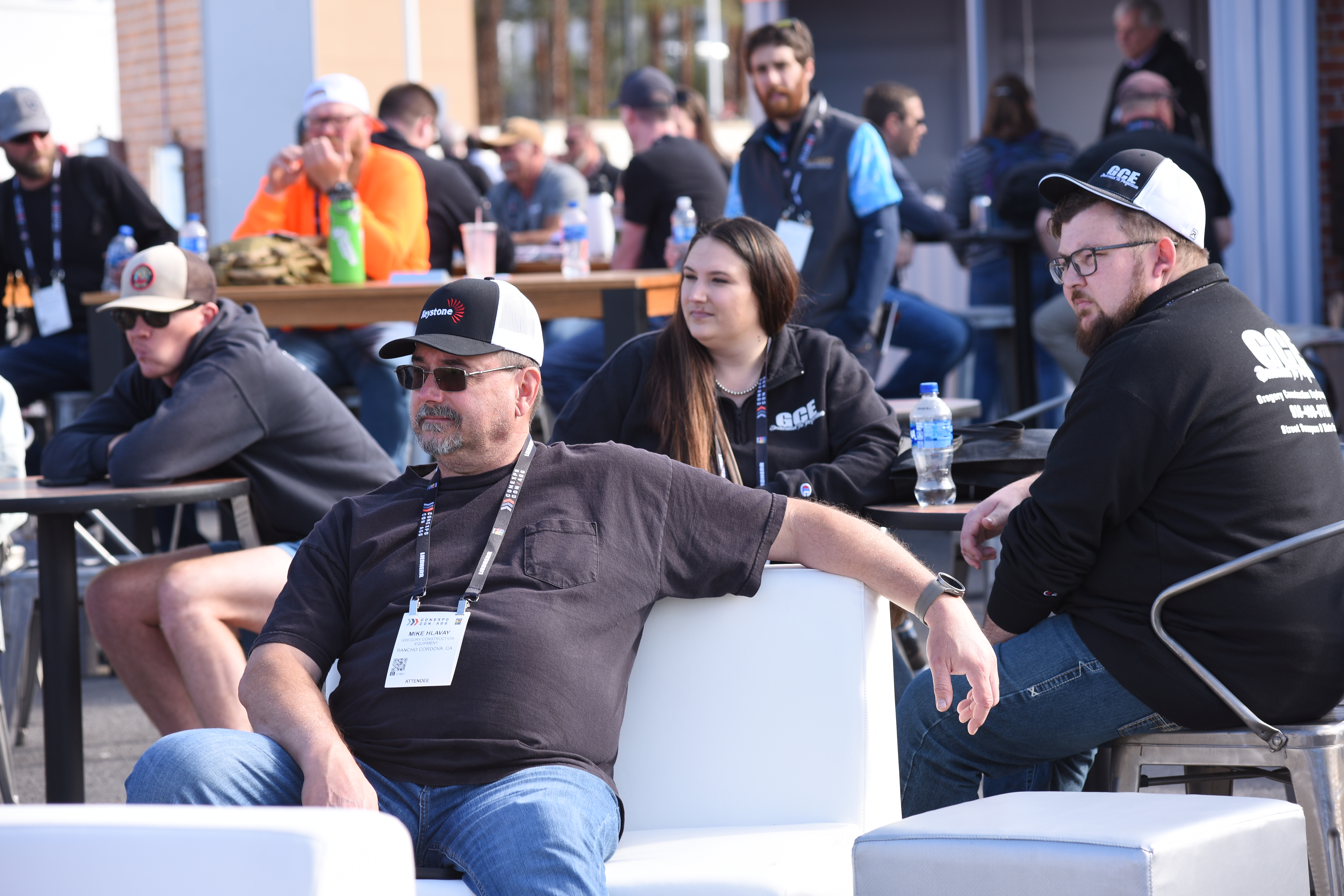 People seated outdoors at tables at the Community Zone, socializing and wearing lanyards and hats, with beverages on the tables and a crowd in the background